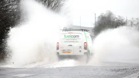 A white van drives through a large puddle in the road. Spray comes off each side of the vehicle. To the right of it the spray is vehicle high. To the left the spray is twice the height of the vehicle. There is a leafless tree to the left of the van.
