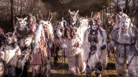 Richard Townsley The Krampus wear elaborate, demonic-inspired animal costumes. Here they are at a previous event walking through a sun-dappled forest, creating an eerie scene. There are six of them in white in this image.