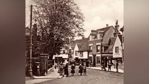 Kevin Fuller c/o Stephen Nunn A black and white photo shows buildings with dormer windows and a wide street outside. A tree is opposite the buildings and next to the tree is a large, ornate pipe.