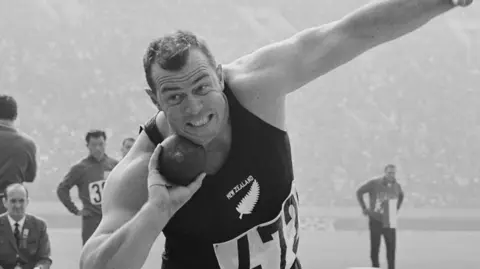 Getty Images A black-and-white image of a shot-putter poised about to launch the putt - he has one arm holding the ball towards his face and the other arm out. He is wearing a black New Zealand bib with the number 472 - officials and a large stadium crowd are seen behind him 