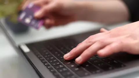 PA Media A woman's hands at the keyboard of a black laptop computer. Out of focus, she is holding a bank card in her right hand.