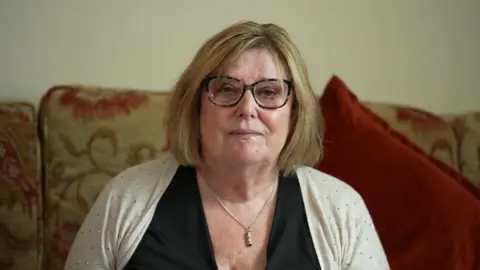 Christine is sat on her sofa wearing a cream cardigan and looks sad