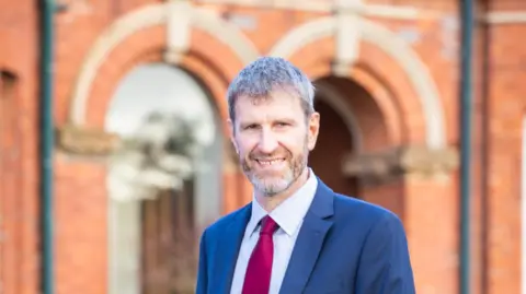 Friends' School Lisburn A man with short grey hair wears a navy suit and blue shirt with a red tie.
