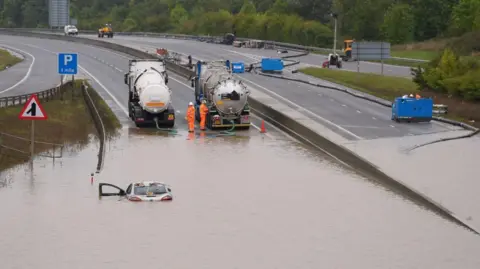 PA Media A car submerged in the floodwater covering the dual carriageway, with pipes trailing from two tankers and two workmen standing by