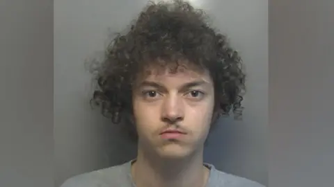 Hertfordshire Constabulary Kierson Murray mugshot wearing a grey t-shirt. He has brown eyes and very curly bushy hair. He  is starting at the camera. He has a light moustache.