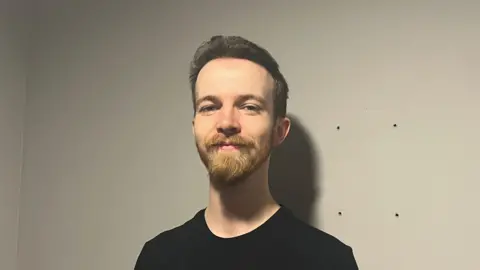 Isabella Verona/BBC Matthew Cook standing indoors against a plain light-coloured wall. The person is wearing a black short-sleeve T‑shirt and has short hair and facial hair.