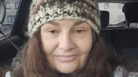 Diane Barnes A middle-aged white woman sitting in a car and taking a selfie. She has dark hair and is wearing a knitted hat. There are houses in the distance. 