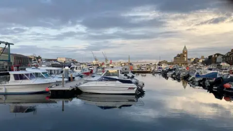 BBC An image of white boats moored in rows St Sampson's harbour in Guernsey. The water is still and reflecting a cloudy sky, in the background is a clock tower and cranes, as well as brown building. 