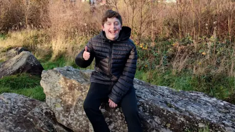 Family photo 13-year-old Kyle sitting on a rock overlooking his hometown, Pontypridd. He has short black hair and is wearing a black puffer coat. He is smiling and making a thumbs up gesture.