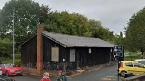 A dark building in a car park with a grey roof and a brick chimney