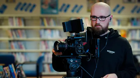 Callum McCourt, TMM Productions Thomas is adjusting a black professional camera. He's wearing dark rimmed glasses and a black hoody. The blurred background shows library shelves.