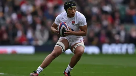 Getty Images A female rugby player holding the ball about to run. She is wearing a white England kit and a black scrum cap on her head. She is looking to the side but there are no other players in the picture.