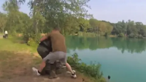 A man in grey shorts and a brown top wrestling a man wearing green trousers and a dark top, by the side of a lake. The image is blurred.