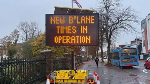 A digital sign that states new b'lane times in operation. Oange and white cones are around the sign. A yellow frame is holding it up. A union flag is in the background. There's a blue bus on the bus lane and cars on the other lanes. It's a wet day with puddles in the street. 