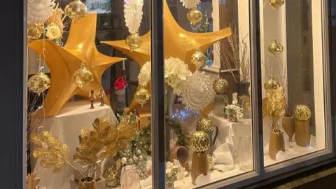 A Christmas window display at a florist, featuring decorations, wreathes, white paper snowflakes and large gold stars.