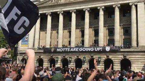 Hundreds of people clapping towards a council house balcony with a banner that reads NOTTS ARE GOING UP