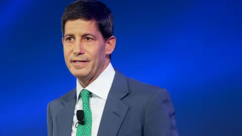 Head and shoulders of Kevin Warsh against a blue background. He is wearing a greyish blue suit with a bright green tie and white shirt.