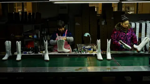 Xiqing Wang / BBC Two female workers making white cowboy boots, which are sitting on a green travellator. The women are dressed warmly in coats and thick jumpers, while most of the factory behind them appears to be in darkness