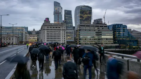 Getty Images Blurred motion view of commuters with umbrellas in London going to work on a rainy, gray day in front of the city skyline during morning rush hour