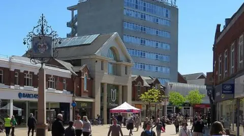 John Sutton/Geograph Chelmsford High Street in the sun with a range of shops and lots of people, with an office block in the background.