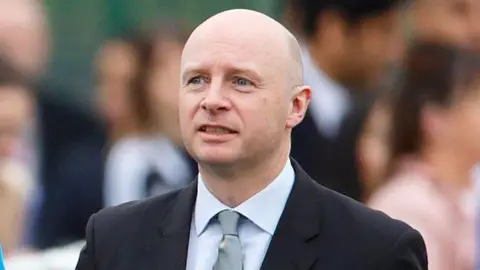 Liam Byrne wearing a suit and shirt and pale blue tie. There are people blurred in the background.