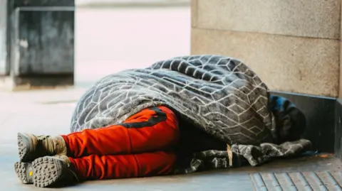 A person with trainers and orange trousers lying underneath a patterned blanket on a paved ground.