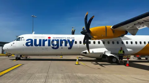 A white airline at Guernsey Airport with the word Aurigny in blue and a red dot over the i. 