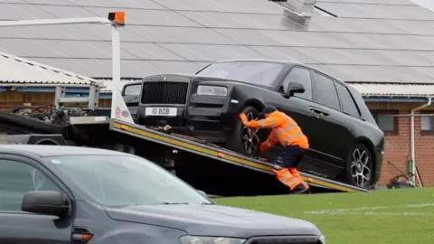 Stuart Woodward/BBC A black Rolls-Royce with the number plate I0 BDB is being secured on to a ramp or a trailer by a man wearing orange high-vis. Behind is a large roof with solar panels on it.