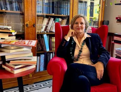 Judge Paola di Nicola sits in an arm chair looking directly into the camera. She is surrounded by books and is wearing a suit and a statement necklace. 