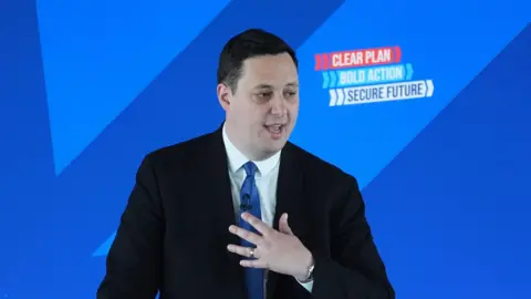 Ben Houchen speaking on stage. He's standing in front of a blue background and is wearing a dark suit and light blue tie.