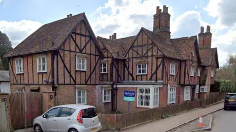 Google The house that was formerly the Barn School in Much Hadham. It is a large two-storey half-timbered red brick building with roofs and gables pointing in different directions. There is a For Sale sign at the front a silver car is parked on the driveway.