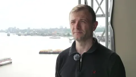 Craig Robinson stands on his balcony. Scaffolding can be seen behind him 
