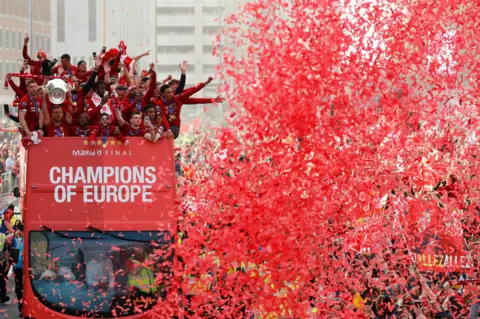 Red confetti in the air in front of the Liverpool bus in 2019. Players raise their arms in celebration on the open-top bus.