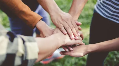 A group of people putting their hands on top of each other. They are standing on grass.
