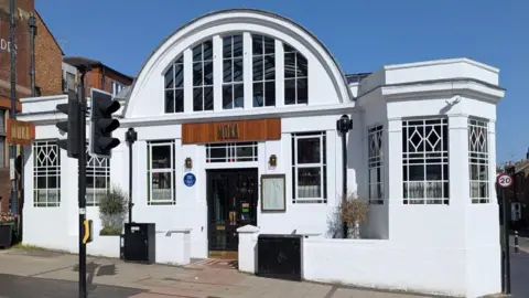 Tim Boatswain A blue plaque honouring Samuel Ryder is attached to a white building that is now a restaurant in St Albans. The former Seed and Exhibition Hall is an Art Deco style building, with lots of windows, and has a restaurant sign above its black doors.