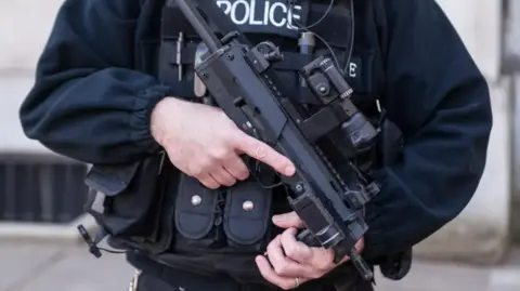 A close up image of an armed police officer holding a gun. The image is just of their chest and the gun. 