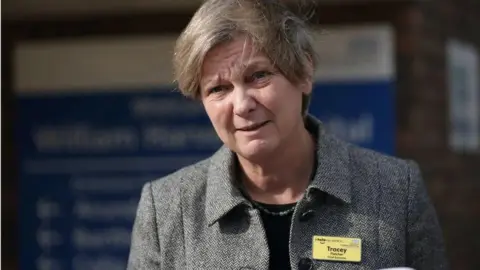 A blonde woman in a grey jacket and a yellow name tag with the name "Tracey" written on it.