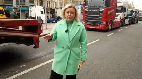 Sara Girvin in a green coat stood in a road next to stopped lorries