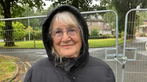 Campaigner Denise Major wearing a black coat with the hood up, smiling in front of a gate by the Dilke Memorial Hospital.