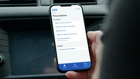 NHS A still taken from the campaign video showing a person sitting in a car with a dashboard in the background. They are holding their phone up in their right hand, and the screen shows they are using the NHS App. They are paused on a page about ordering prescriptions. 