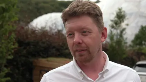A man wearing a white shirt speaks to a reporter.