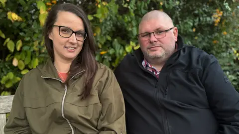 Zoe and Lewis Steeper sat on Oliver's bench in a park. They are both wearing jackets and sat in front of a green hedge with some yellow leaves.