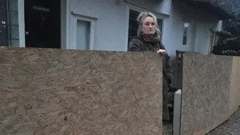 Henrietta Edwards Blonde woman standing next to wooden boards while standing outside her cottage in Essex