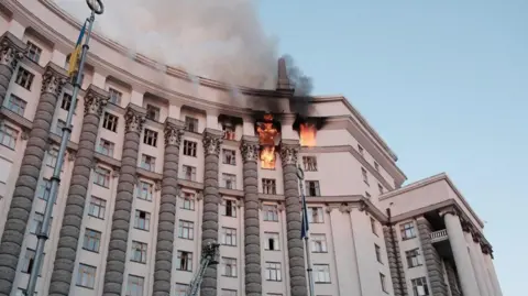Telegram/svyrydenkoy The image shows a large grey and light stone building, the main Ukraine government building, set against a blue sky with eight windows wide and six to seven windows vertical, with the top two floors on the right hand side on fire. There are cranes attempting to extinguish the fire,
