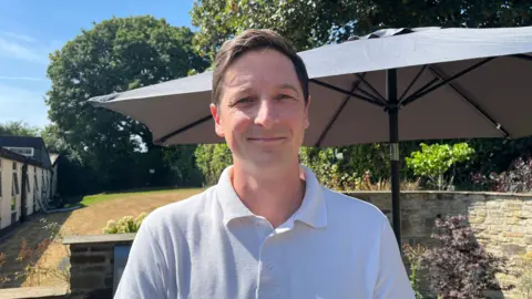 Ellen Knight/BBC Photo of Klee, who is looking into the camera and smiling. He is wearing a white polo shirt, and is stood in front of a large parasol in the hospital garden. The garden is surrounded by low stone walls, which have plants and flowers growing up them. The sky is blue and there is strong sunshine. 