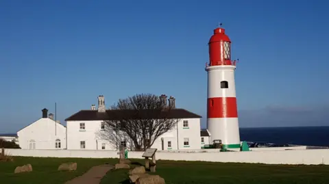 The Souter Lighthouse is a large two-storey white house adjoining a red and white striped lighthouse with large lantern at the top, the outside of which is all red. It is a clear blue day with a calm sea in the distance.