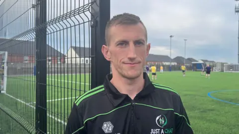 A man wearing a black and green "Just a Chat" training top is standing beside a fence of an outdoor football pitch, looking at the camera. Players are training in the background beneath an overcast sky
