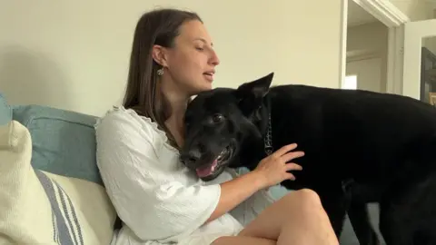 K-Jo sits on a sofa, cuddling a large, black dog. She wears a white summer dress.