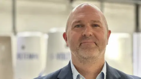 A man with a bald head and a faint beard with a grey jacket and white shirt in front of a blurred background