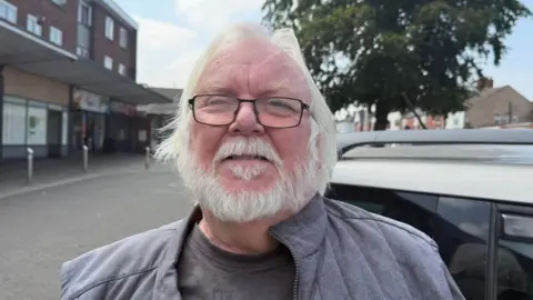 The photo shows Rob Barnard looking at the camera. He has white hair and a white beard, and is wearing black rimmed glasses. He's also wearing a grey jacket. He's standing in front of a car. Behind him, to his left, there are some shops. 